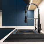 Matte black spring faucet above a deep sink and roll up rack, blue fronts and a marble backsplash under LED lighting, no people, neat contemporary space.