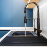 Low angle view of a professional spring faucet with running water, a black sink and roll up drainer. Blue handleless cabinetry and LED shelf light deliver a premium vibe.